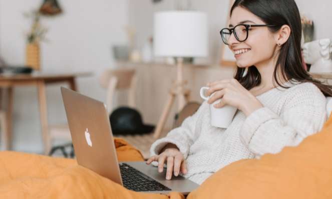 Girl smiling while working on her laptop and holding a cup of coffee. She is seated on her couch and wearing glasses.