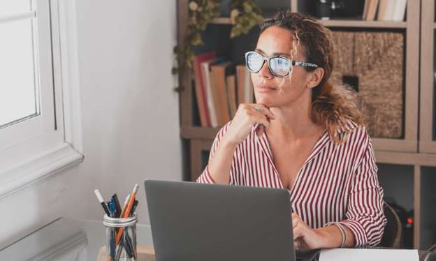 A woman sitting at a desk with one hand under her chin and the other resting on the laptop keyboard. There is a jar of pens and paper on the desk. 