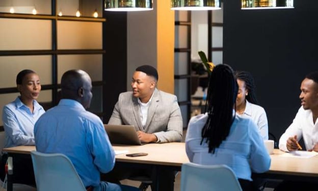 A group of people are sitting around a large conference table, engaged in a conversation. One man has a laptop, and another man has a notepad.