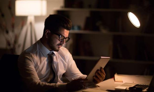 Man seated at a desk in a dark room, looking at an iPad. There is a lamp on the desk as well as one in the background. He's wearing glasses and a tie.