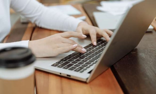 A woman is sitting at a desk with her hands typing on the keyboard of an open laptop. There is also a coffee cup and notebook on the table. 