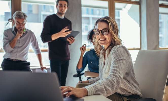 A team is working in a meeting room, two men, two women. One woman is in the foreground using a laptop, wearing glasses, and smiling.