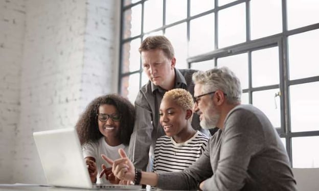 Three people are sitting at a desk with one person standing. All four are looking at a laptop screen while smiling. 