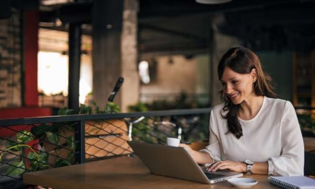 A woman sitting at a table outside at night, working on a laptop, smiling. On the table is a saucer and a notebook. 