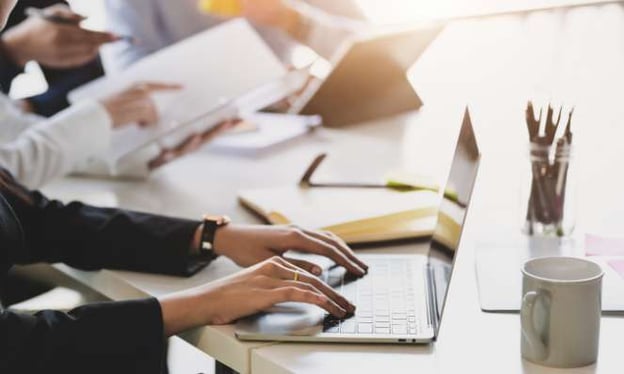 A woman sitting at a desk with her hands resting on a laptop's keyboard. In the background are three others reviewing notes and a tablet. 