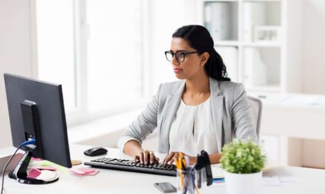 Woman wearing glasses and a gray blazer, typing on a keyboard and looking at a computer monitor while seated at her desk.