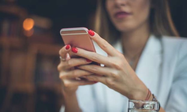 A woman is looking at her mobile phone. It is arose gold phone and her nails are painted red. 