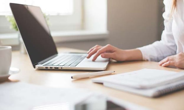 A person sits at a desk with their hand on a laptop keyboard. A marker, notebook, and mug are also on the table. 