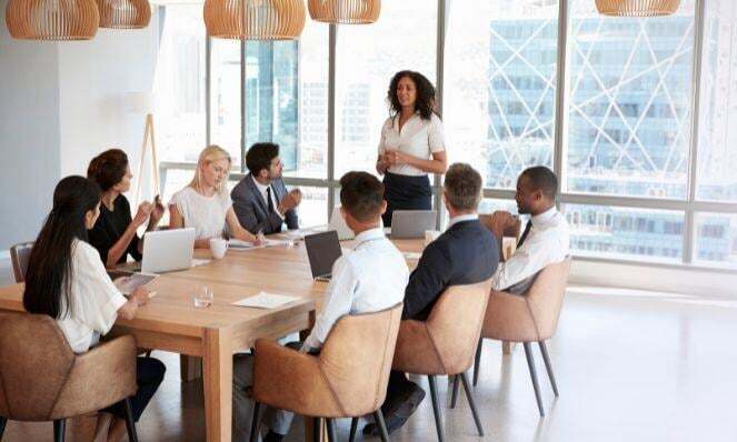 A team of coworkers seated around a conference table as one woman stands and speaks to the group. There is a large window behind her.