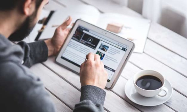 A man with a beard is seated at a table, reading a newsletter on a tablet in his hand. A magazine is in the background and a coffee cup next to him.