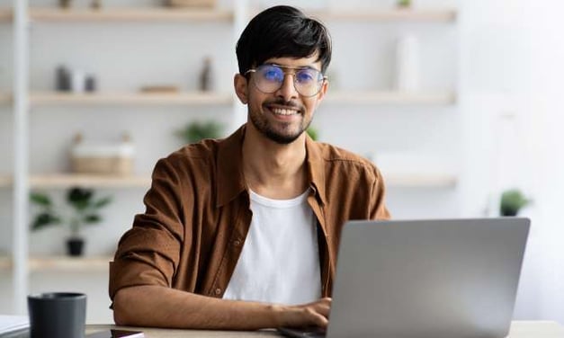 A man sitting at a table while on a laptop looking up and smiling. There is a cell phone and mug on the table. He has large circular glasses.