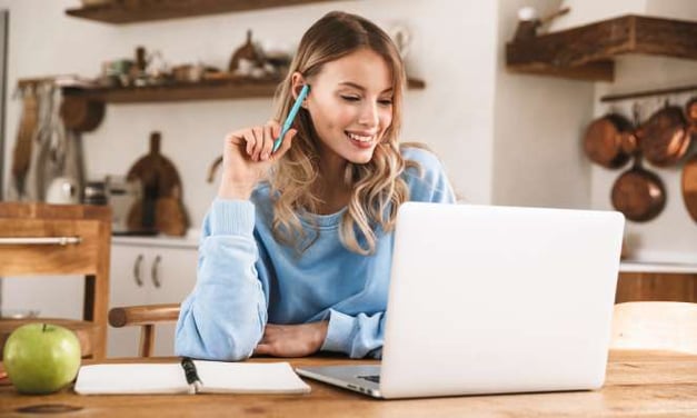 A woman sits at a desk looking at a laptop screen with a notebook and apple at her side. She is smiling and holding a blue pen. 
