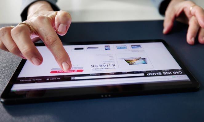 Image of a person using a tablet placed on a tabletop to do online shopping. They are pressing the purchse button with one hand and the other is on the table.