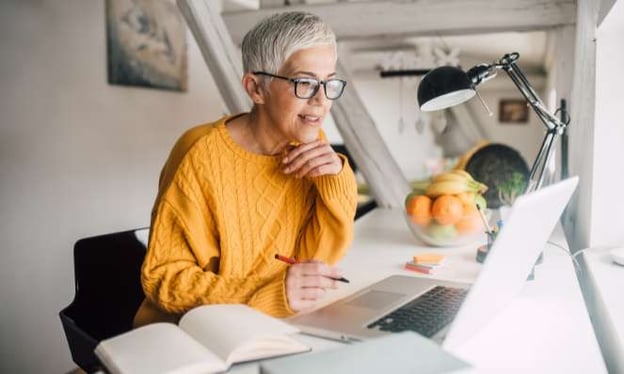A lady in an orange sweater sits at a desk, looking at a laptop screen. A lamp, an open book, and a bowl of fruit are on the desk.