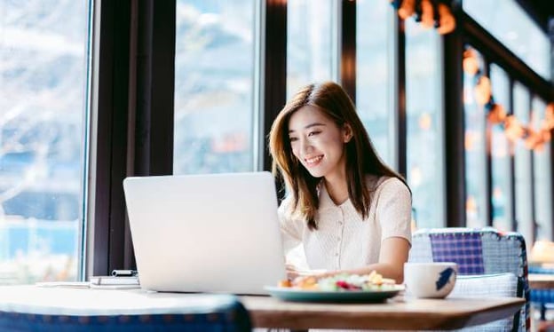 A woman sitting at a table in a cafe working on her laptop and smiling. On the table is a mug and a plate of fruit. 