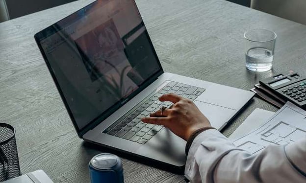 A person working on a laptop on a desk. There is a hand on the keyboard. A glass of water, a calculator, and papers are on the desk. 