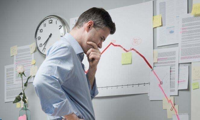 Person thinking intensely of a solution to a problem while standing in front of a wall with reports and graphs on it. There is also a clock.