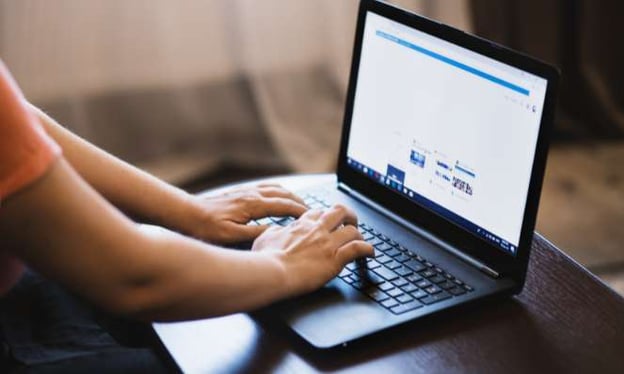 A person rests their hands on a laptop keyboard that sits on a table. The laptop screen has open articles on it.