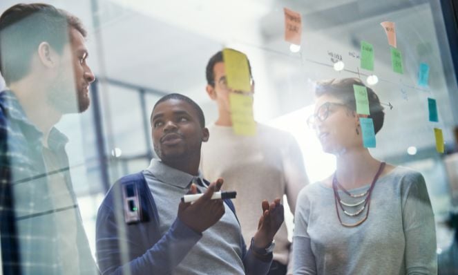 A group of multicultural coworkers working in front of a glass window with sticky notes on it, discussing business topics.