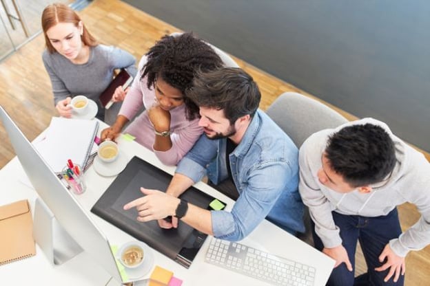 Four people seated at a desk, looking at a computer monitor. One person is pointing at the screen. Photo is taken from above.