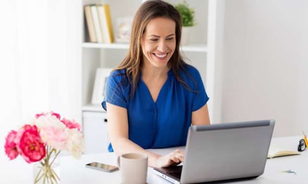 A woman is sitting at a desk, smiling while working on a laptop. A vase with pink flowers, a cell phone, a mug, and a notebook is on the desk. 
