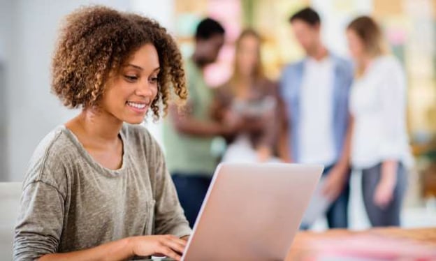 A woman sitting at a desk, smiling, and working on a laptop. She has curly brown hair and is wearing a loose-fitted grey shirt. 