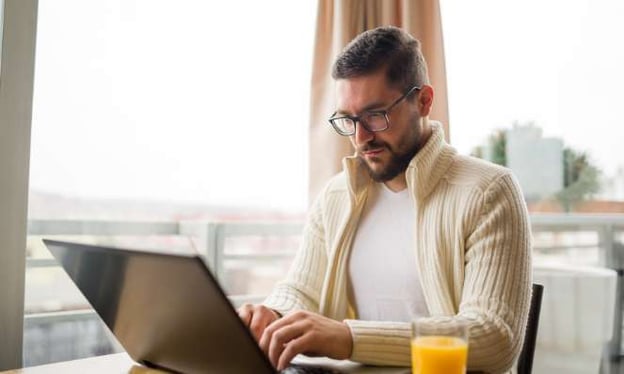 A man wearing glasses and a cream-colored sweater is sitting at a desk working on a  laptop. On the table is a glass of orange juice.
