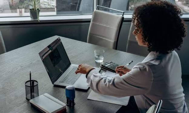 A woman is sitting at a table working on a laptop. She is in a dress shirt. A glass of water, a calculator, paper, and a notebook are on the table.