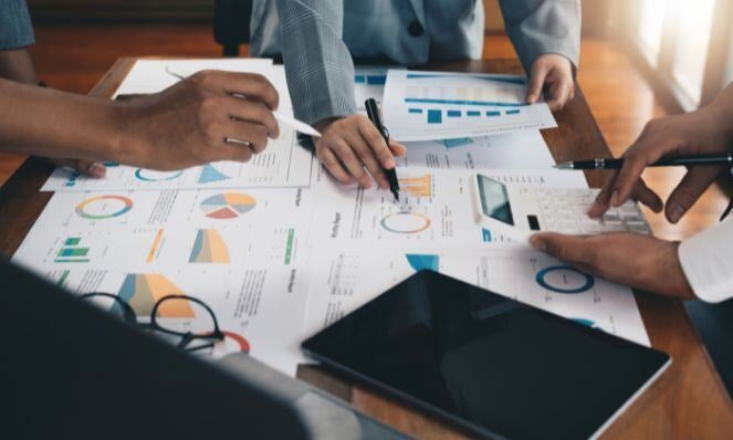 Agency employees reviewing papers with many reports on them. One person is holding a calculator while the other two hold pens.