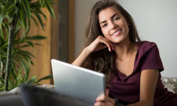 A woman sitting with a laptop on her lap while smiling and looking at the camera. She is a brunette with long hair and a purple t-shirt.