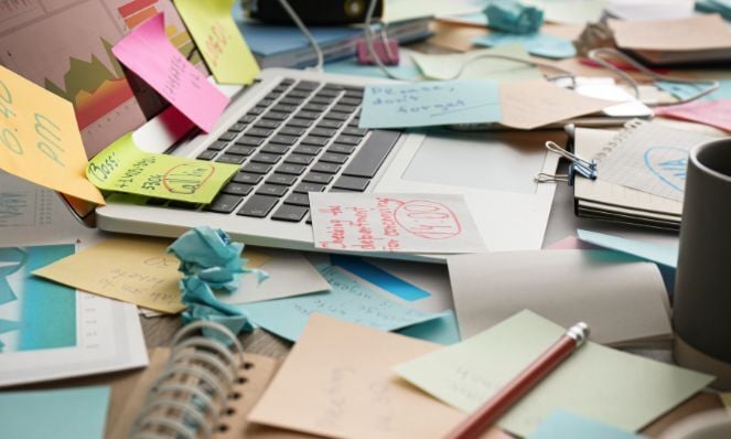 A photo of a very cluttered desk with many sticky notes, note pads, and a laptop. The desk itself is hardly visible. There is a pencil and a coffee mug, as well.