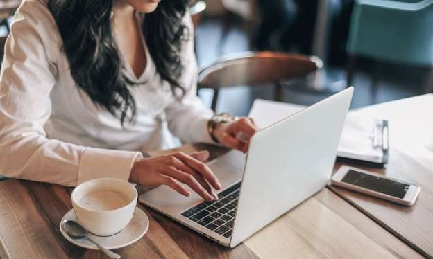 A woman sitting at a table working on a laptop. On the table is a coffee cup on a saucer with a spoon, a cell phone, and a notebook. 