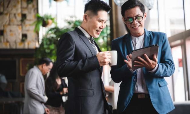 Two men, standing, one holding coffee, other holding tablet which they are both looking at and smiling, in an office, windows and a man in background.