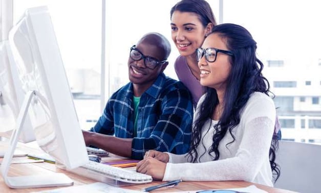 Three people smiling looking at a desktop monitor while 2 of them are sitting at the desk and 1 of them is standing. 2 female and 1 male.