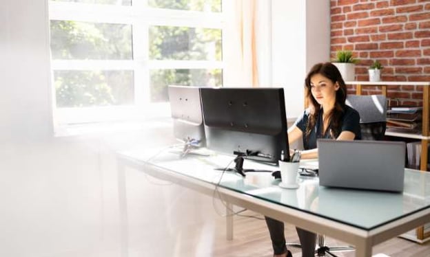 A woman is working while sitting at a desk. She has a laptop and two computer monitors in front of her. There is a cup with pens on the desk.