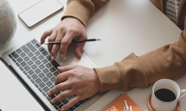 Hands typing on a laptop keyboard; one holds a pencil. A cell phone, desk, notepad, AirPods, and a coffee cup are on the table.