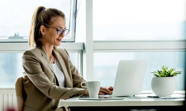 A woman wearing glasses sits at a desk, working on a laptop. A tablet, coffee cup, and plant are on the desk. There are windows behind her.