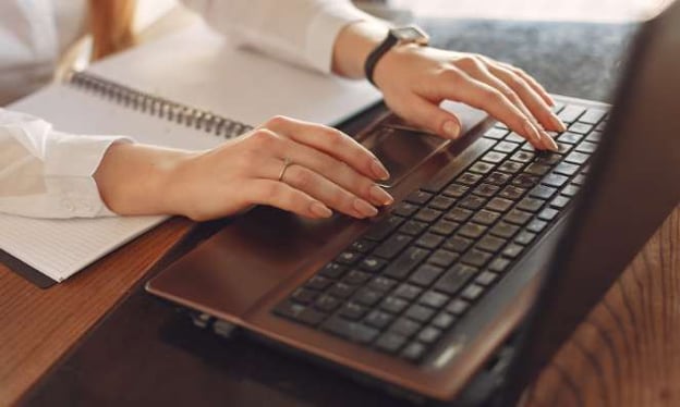 A woman's hands are resting on the keyboard of an open laptop while sitting at a desk. On the desk next to the laptop is a notebook. 