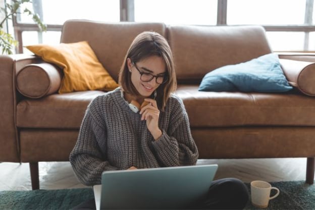 Woman seated on the floor with a laptop, she is wearing a brown sweater and glasses. A brown couch is behind her and a white coffee mug next to her.