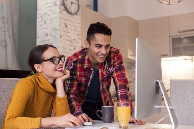 Two people at a desk, one seated, one standing, looking at a computer monitor. A glass of orange juice and a cup of coffee are on the desk, too.