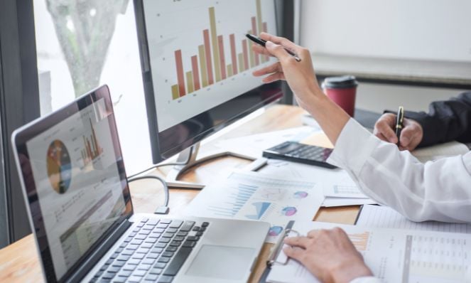 Photo of two people whose arms and hands are only visible. They are seated at a desk together and looking at a laptop and monitor, both with pie charts and bar graphs on them. There are also many papers on the desk which also have reports on them.