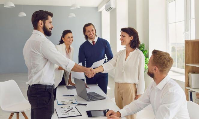 Professionals shaking hands and smiling while standing around a conference table while workmates look on and smile, as well.