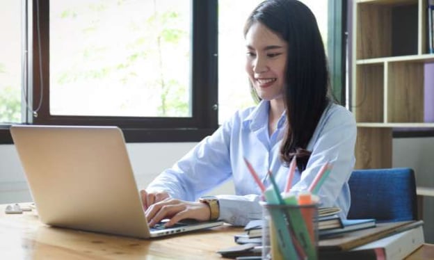 A woman smiling while working at a on a laptop on a desk. The desk has a container of markers and a stack of books on it. A window is beside her.