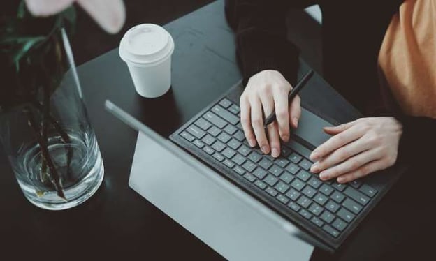 A woman sitting at a desk with her hands resting on the keyboard of an open laptop. On the desk is a coffee cup and a vase with flowers. 