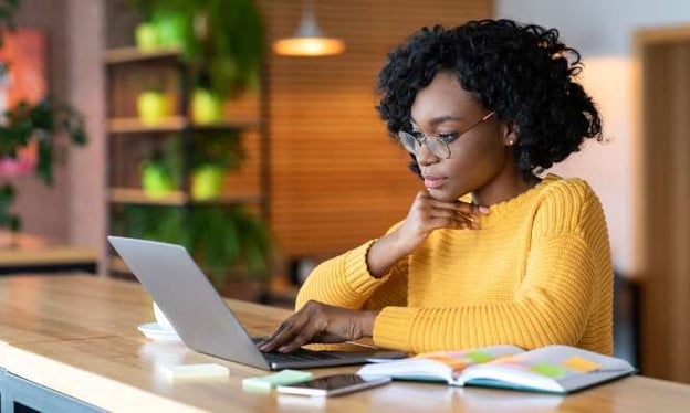 A woman wearing glasses with her chin resting on her hand looks at a laptop screen while sitting at a table. There is a notebook on the table. 