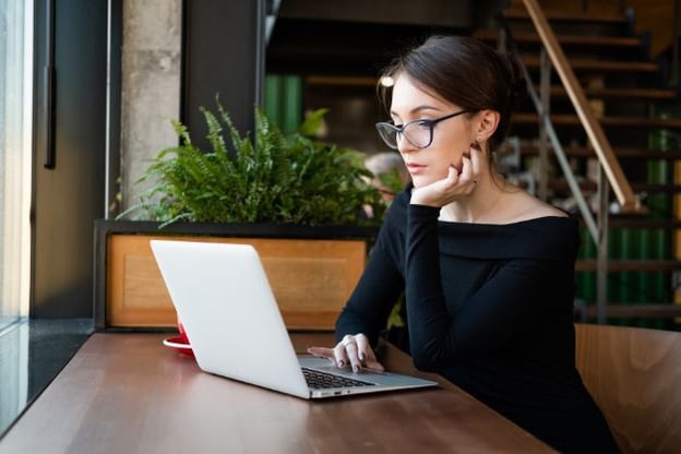 Woman wearing glasses is seated at a desk with her laptop open, one hand under her chin, the other on the track pad. Potted plants behind her.
