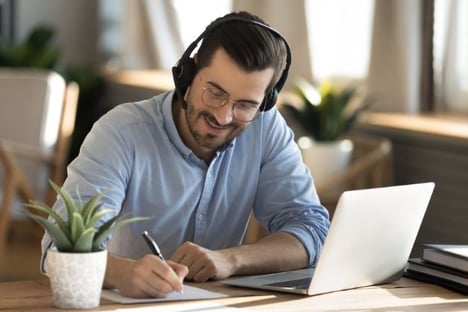 Man wearing glasses and headphones, seated at desk, laptop open in front of him, he is writing with a pen on paper. Potted plant in the foreground.