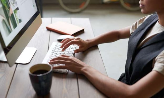 A woman sits at a desk and is looking at a monitor and typing on a keyboard. On the monitor are images. On the desk is a book and a coffee cup.
