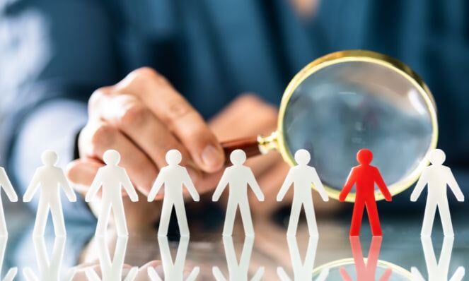 Person looking into a magnifying glass at a cutout figure of a person in red surrounded by cutouts of people in white in a line.