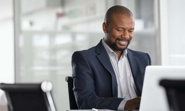 A man sitting at a desk dressed in a suit, smiling, looking at a laptop screen. He is wearing a white dress shirt with a navy blazer overtop. 